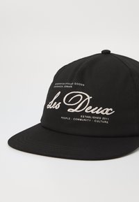 Black cap made of cotton, featuring embroidered white text "Les Deux" and additional text. Designed with a curved brim and six-panel structure.