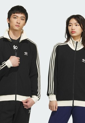 Zwarte geribbelde jas met rits, witte accenten en drie strepen op de mouwen; heeft een hoge kraag, logo op de borst en aansluitende manchetten.