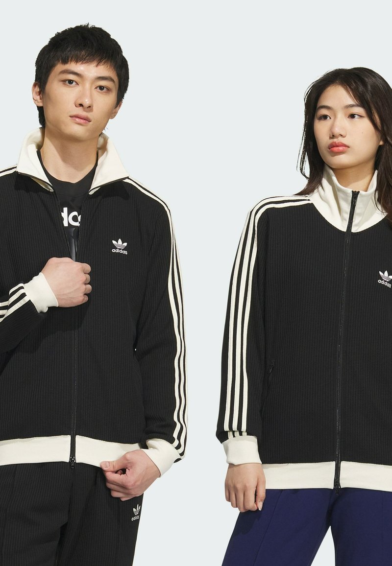 Chaqueta negra de cremallera con textura acanalada, acentos blancos y tres rayas en las mangas; cuenta con cuello alto, logo en el pecho y puños ajustados.