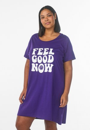 Kobieta stoi i uśmiecha się, ubrana w fioletową sukienkę typu t-shirt z wyraźnym białym napisem „"FEEL GOOD NOW"” i małą uśmiechniętą buźką.