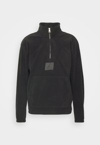 Pullover in pile nero con colletto alto, zip a quarti sul davanti e tasca frontale. Caratterizzato da una toppa con logo sul petto. Tessuto morbido.