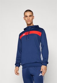 Veste à capuche zippée bleu marine avec une bande horizontale rouge, présentant des bandes latérales blanches et le logo Adidas sur la poitrine. Tissu lisse et léger.