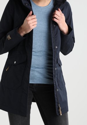 Persona que lleva un abrigo azul marino abotonado y con cremallera sobre una camisa azul claro con textura y vaqueros oscuros, ajustándose el cuello del abrigo con ambas manos.