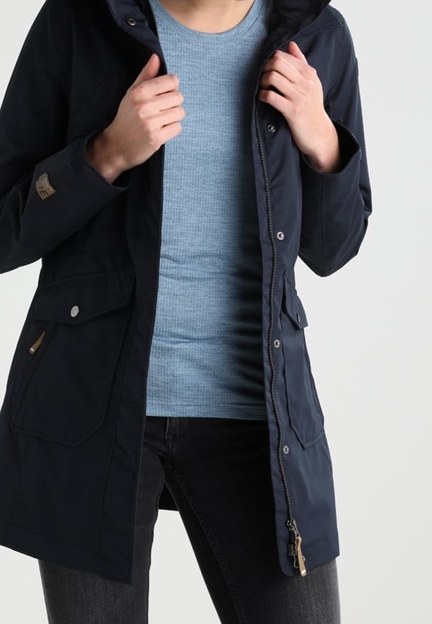 Personne portant un manteau bleu marine boutonné et zippé par-dessus une chemise bleu clair texturée et un jean foncé, ajustant le col du manteau avec les deux mains.