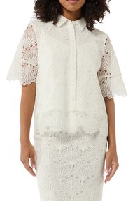 Weiße Spitzenbluse mit kurzen Ärmeln, Knopfverschluss und zartem Blumenmuster. Der Stoff hat ein strukturiertes, schichtiertes Aussehen.