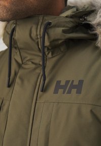 Maslinasto zelena puffa jakna s crnim logotipom, podesivim vezicama i mekanim krznom oko vrata. Ima džepove na zatvarač i teksturiranu površinu.