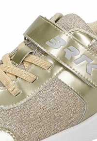Buty sportowe z jasnosrebrzystym metalowym paskiem i błyszczącą beżową tkaniną na górze. Posiada beżowe sznurowadła i gumową podeszwę. Srebrne logo na pasku.