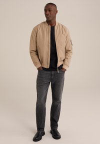 Beige bombr jacket s předním zipem, žebrovanými manžetami a bočními kapsami. Spárován s tmavými džíny a černými botami, stojící na neutrálním pozadí.