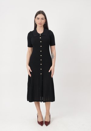 Femme debout face caméra portant une robe midi noire côtelée à manches courtes, boutons dorés, un col et des talons rouges pointus.