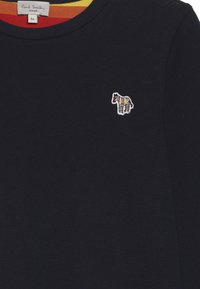 Námořnická modrá bavlněná košile s dlouhým rukávem, kulatým výstřihem a malým výšivkou zebry v barevných pruzích na levé části hrudi.