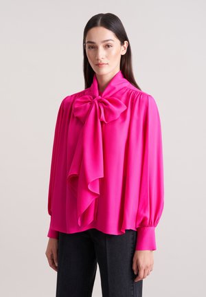 Fuchsia Bluse mit einer großen Schleife am Kragen, langen, gepufften Ärmeln und einem fließenden Design. Glattes Material mit einem leicht glänzenden Finish.