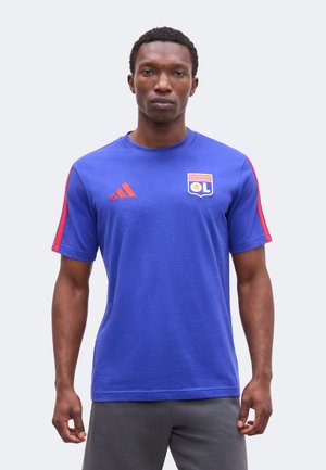 Bărbat purtând un tricou albastru de fotbal Olympique Lyonnais cu dungi roșii Adidas pe mâneci și pantaloni gri, stând într-un fundal simplu.