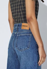 Jeans en denim bleu taille haute avec une finition texturée, dotés de poches arrière et d'une étiquette en cuir beige de marque sur la ceinture.