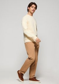 Sudadera de crema clara con cuello redondo, combinada con pantalones beige ajustados y zapatillas marrones. El modelo camina frente a un fondo gris liso.