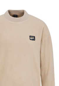 Felpa in pile beige con colletto alto, caratterizzata da un'etichetta logo rettangolare nera che mostra montagne e strisce sul petto sinistro.