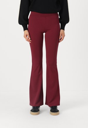 Person iført tætsiddende, bordeauxfarvede flare-bukser med sideslids og sorte sneakers mod en ensfarvet hvid baggrund.