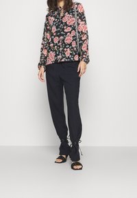 Blouse noire à motifs floraux avec des roses, manches longues et encolure fendue, associée à un pantalon ample bleu marine et des sandales à lanières noires.