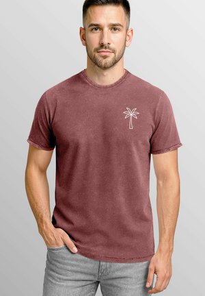 Bordeauxrotes T-Shirt aus einem weichen, strukturierten Stoff mit kurzen Ärmeln. Mit einem weißen Palmenbaum-Design auf der linken Brust. Stylisch kombiniert mit grauen Jeans.