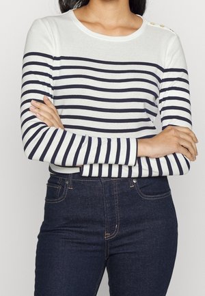 Femme portant une chemise blanche à manches longues avec des rayures horizontales bleu marine et des boutons dorés sur l'épaule, accompagnée d'un jean bleu foncé.