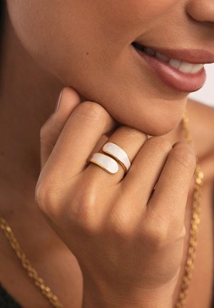 Anillos con acentos dorados y mármol blanco en un diseño curvado de doble banda, llevados en una mano con un acabado de piel texturizada.