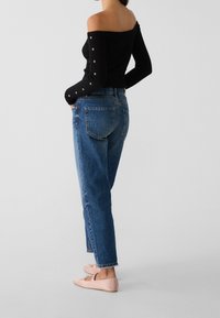 Svart off-shoulder topp med ribbad textur, blå högmidjade jeans och blekrosa ballerina-skor med en rosett, sedd bakifrån.