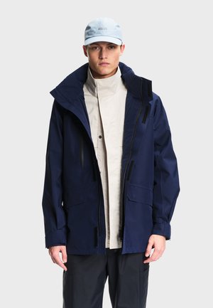 Homme portant une veste bleu marine, une chemise beige à col montant, un pantalon noir et une casquette bleu clair sur un fond uni.