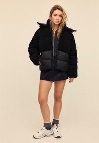 Črna puhasta jakna z zgornjim delom iz teksturirane flisa in spodnjim delom iz podložene najlonske tkanine. Opremljena je z kapuco in sproščenim krojem, kombinirana s črtasto obleko in supergami.