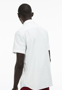 Biała koszula z krótkim rękawem, wykonana z gładkiej bawełny; ma klasyczny kołnierzyk oraz zaokrąglony dół z małym niebieskim akcentem po boku.