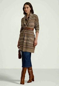 Mulher a usar um cardigã bege estampado com cinto, uma saia de ganga escura, botas castanhas de salto, e a segurar uma pequena bolsa castanha.