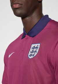 Bordowa koszulka polo z granatowym kołnierzykiem, z białym logo Nike oraz haftowanym herbem Anglii na piersi. Gładka tekstura materiału.