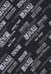 Černá tkanina s opakujícím se bílým textem, včetně "VERSACE JEANS COUTURE," "SINCE 1988," a "BASED IN MILAN," v různých orientacích.