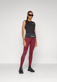 Svart ermeløs topp, røde leggings, grå midje med logo. Svart solbriller, svarte sko. Glatt materiale, tettsittende design.