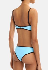 Dwuczęściowy bikini w jasnoniebieskim kolorze z ciemnobrązowymi wykończeniami. Górna część ma cienkie ramiączka i zapięcie z tyłu, natomiast dół charakteryzuje się niskim stanem.