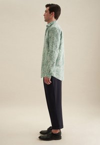 Camicia verde con motivo paisley e colletto button-down, abbinata a pantaloni blu scuro e scarpe nere, vista di lato.