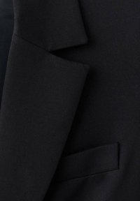 Czarna marynarka garniturowa z teksturowanej tkaniny, ze zbudowanymi ramionami i dyskretną boczną kieszenią. Wyraźne, czyste linie i dopasowany krój.