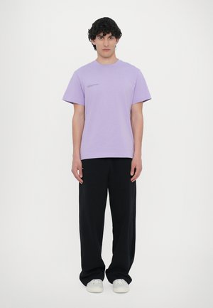Lys lilla T-shirt med rund hals og korte ærmer, parret med sorte vide bukser og hvide sneakers, stående foran en ensfarvet baggrund.