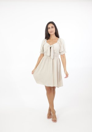 Vrouw glimlacht, draagt een korte beige jurk met pofmouwen en een strik vooraan, staat met één hand de zoom van de jurk vasthoudend, heldere achtergrond.
