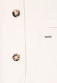 Fehér szövet ferde textúrával, nagy, márványozott gombbal és egy kis márkázott címkével. Egyszerű zsebszerkezet az oldalon.