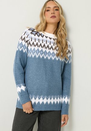 Femme aux longs cheveux blonds portant un pull en maille à motifs bleu et blanc et un pantalon sombre, debout contre un mur clair uni.