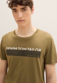 Olívazöld pamut póló kerek nyakkal, fehér nyomott "DENIM TOM TAILOR" logóval és fekete textúrált csíkkal. Sima textúra.