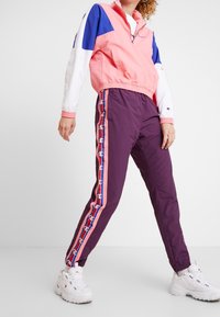Chaqueta rosa y azul con media cremallera y mangas blancas, pantalones morados con rayas laterales rojas y rosas, y zapatillas blancas gruesas.