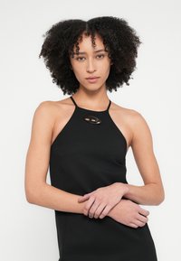 Fekete pánt nélküli ruha, amely kulcslyuk kivágással rendelkezik a nyakkivágásnál, testhez simuló szabással és sima textúrával. A modell összefont karokkal áll.