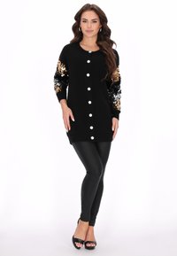 Cardigan negru cu mâneci cu paiete aurii și argintii, cu închidere cu nasturi și lungime mai lungă, asortat cu colanți negri și pantofi cu toc.