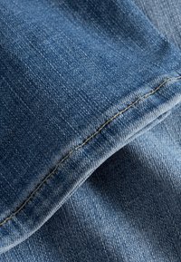 Džínovina ve středně modré barvě, s hladkou texturou, viditelným vzorem tkaní a kontrastním žlutým prošitím podél švů.