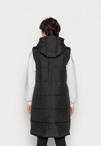 Person med kort mörkt hår iförda en svart ärmlös vadderad huva över en ljusgrå långärmad tröja, vänd bort från kameran mot en enfärgad bakgrund.