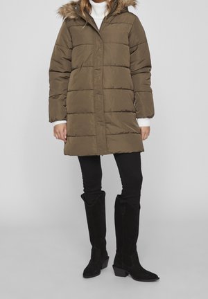 Femme portant un manteau d'hiver matelassé vert olive avec une capuche en fourrure, un pantalon noir et des bottes noires montant aux genoux sur un fond clair uni.
