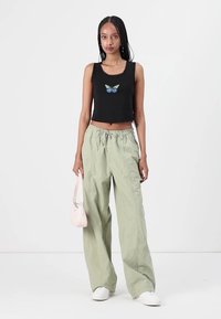 Černý crop top s modrou a zelenou výšivkou motýlů, v kombinaci s volnými, světle zelenými kalhotami a bílými teniskami. Drží malou růžovou tašku.