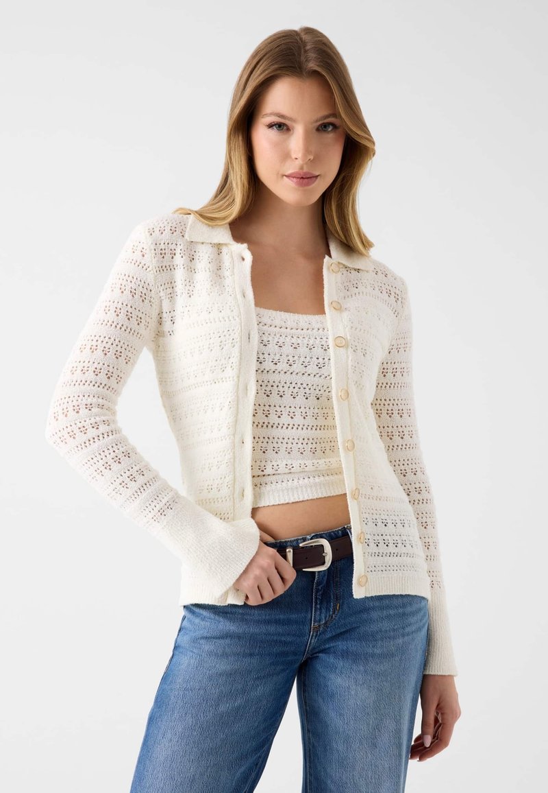 Jeune femme portant un cardigan en crochet blanc et un haut court assorti, associés à un jean bleu et une ceinture marron, posant devant un fond uni.
