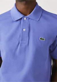 Ljust lila polo skjorta i texturerad tyg, med en treknapplång placket och en grön krokodil-logotyp på vänster bröst.