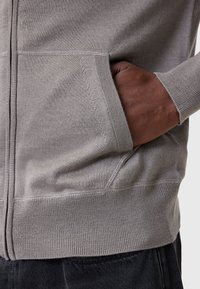 Cardigan grigio in maglia, con zip e tasche laterali. Polsini e fondo a costine con texture liscia, che mette in risalto le cuciture dettagliate.